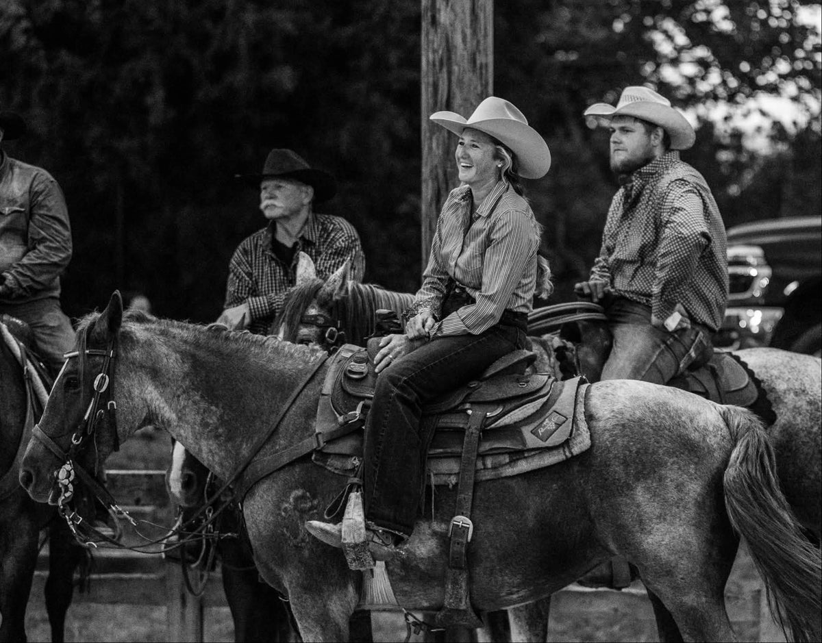 Black and white photograph of a group of cowboys riding horses outdoors, with a woman in a cowboy hat seated on a horse in the center.